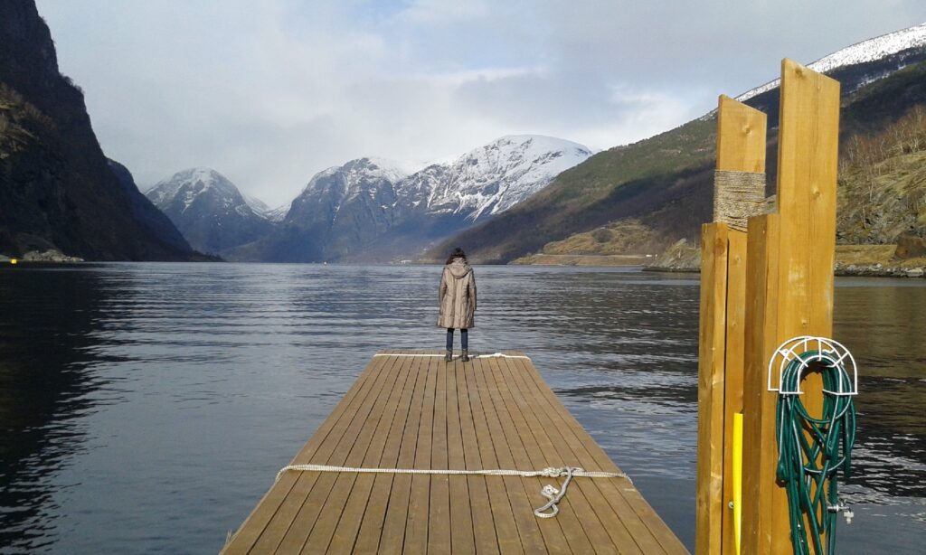 Palno general de los Firodos Noruegos con embarcadero de madera en primer plano y persona de espalda contemplando el paisaje.